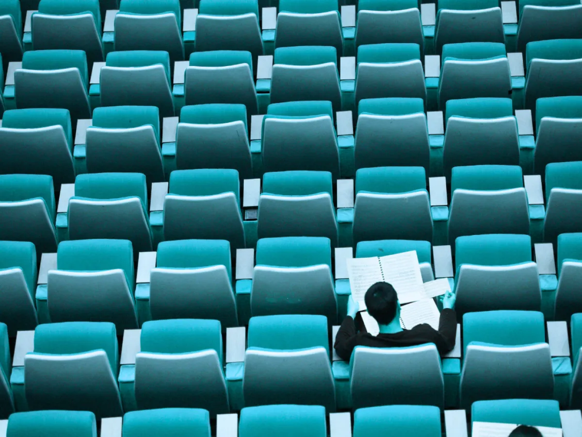 A kép egy magányos férfit ábrázol egy hatalmas, üres nézőtéren vagy előadóteremben. A férfi háttal ül a kamerának, és egy kottát vagy dokumentumot tart a kezében, ami elmélyült figyelmet vagy magányos gyakorlást sugall.